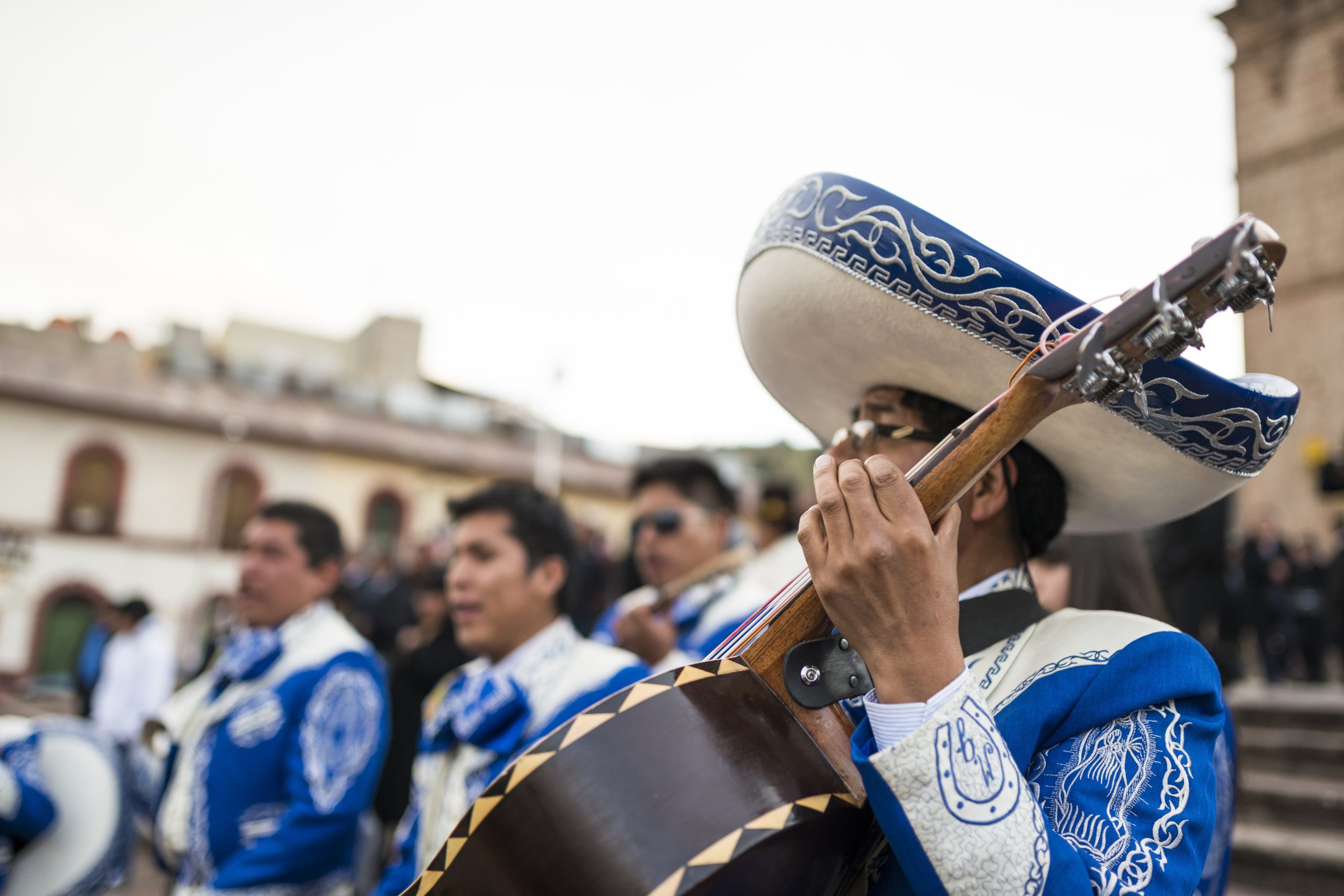 Mariachi musician strumming a guitar with a group of performers in the background.